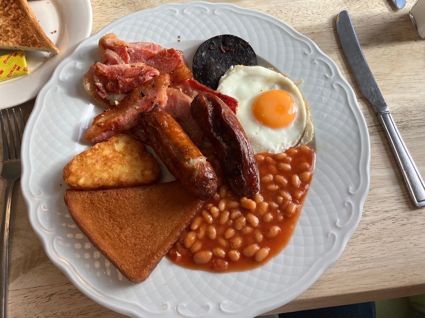 A cooked breakfast on a white plate: bacon, black pudding, a fried egg, sausages, a hash brown, fried bread and beans