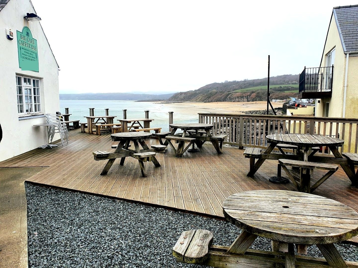 The café's wooden deck with picnic tables, the green Bay Café – Caffi'r Bae sign on the white gable, and Benllech beach and the bay beyond